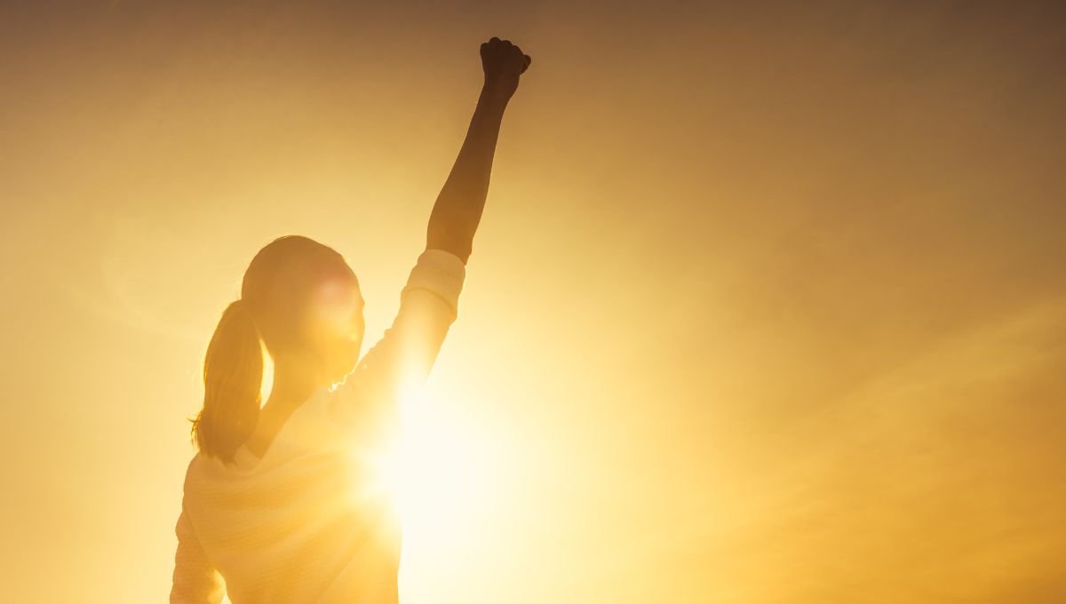 a woman with a pony tail with her fist raised high in victory in front of a sunset